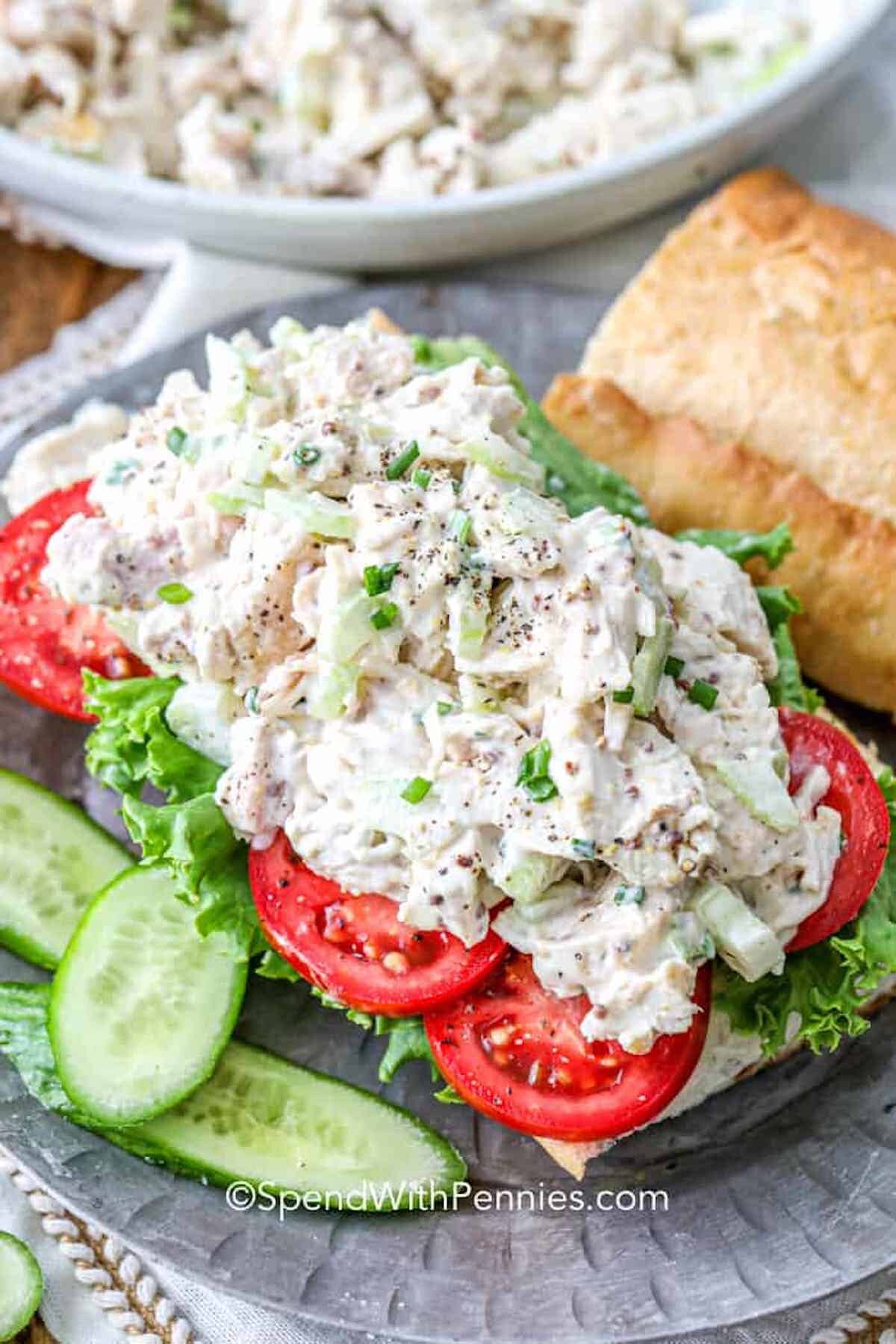 A classic chicken salad on a roll with tomatoes and lettuce next to cucumbers.